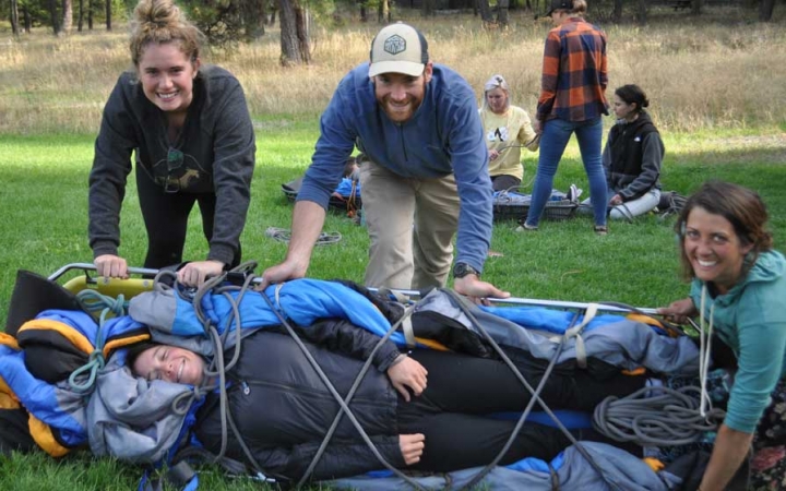 One person lies bound on a stretcher while three people roll them over and smile at the camera. They are all participating in a wilderness first responder course.